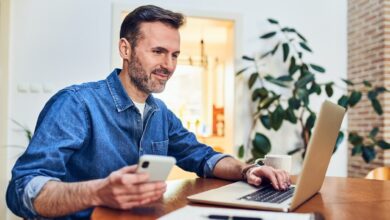 Man sitting at a dining room table looking at his laptop and phone