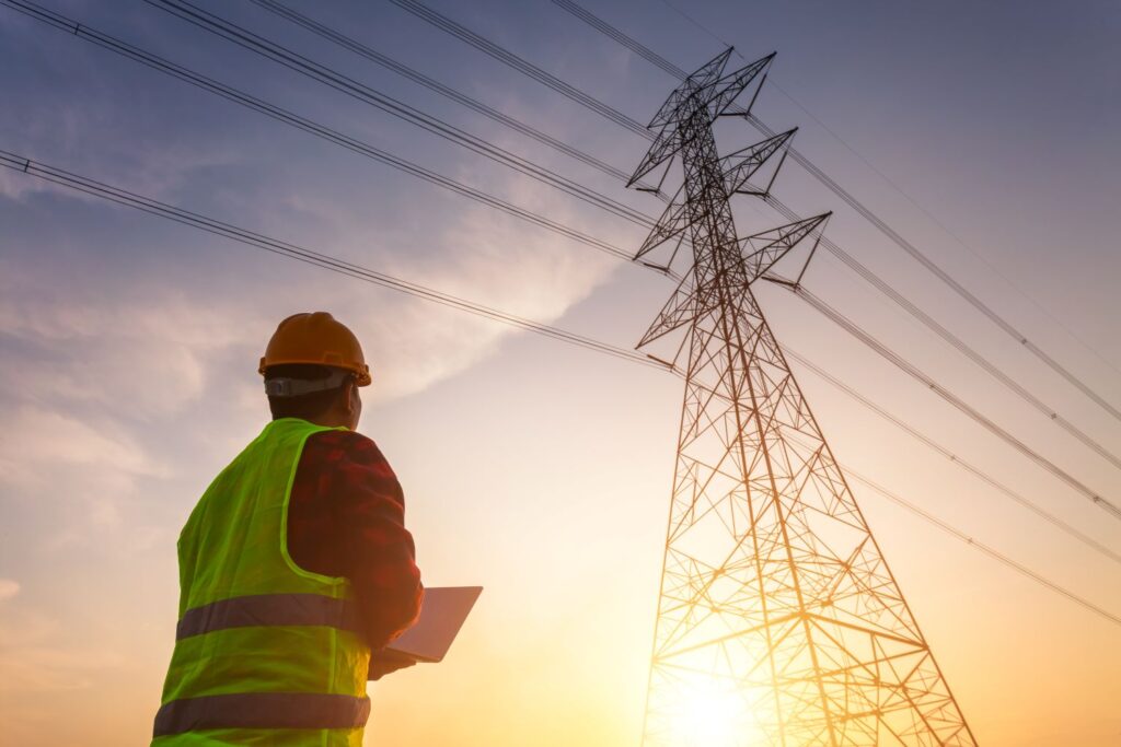 A man in a hard hat holding a laptop as he looks up toward electric transmission line as the sun rises in the background.