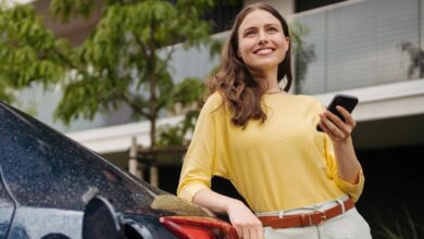 Woman leaning against an electric vehicle while it's charging with a phone in her hand.