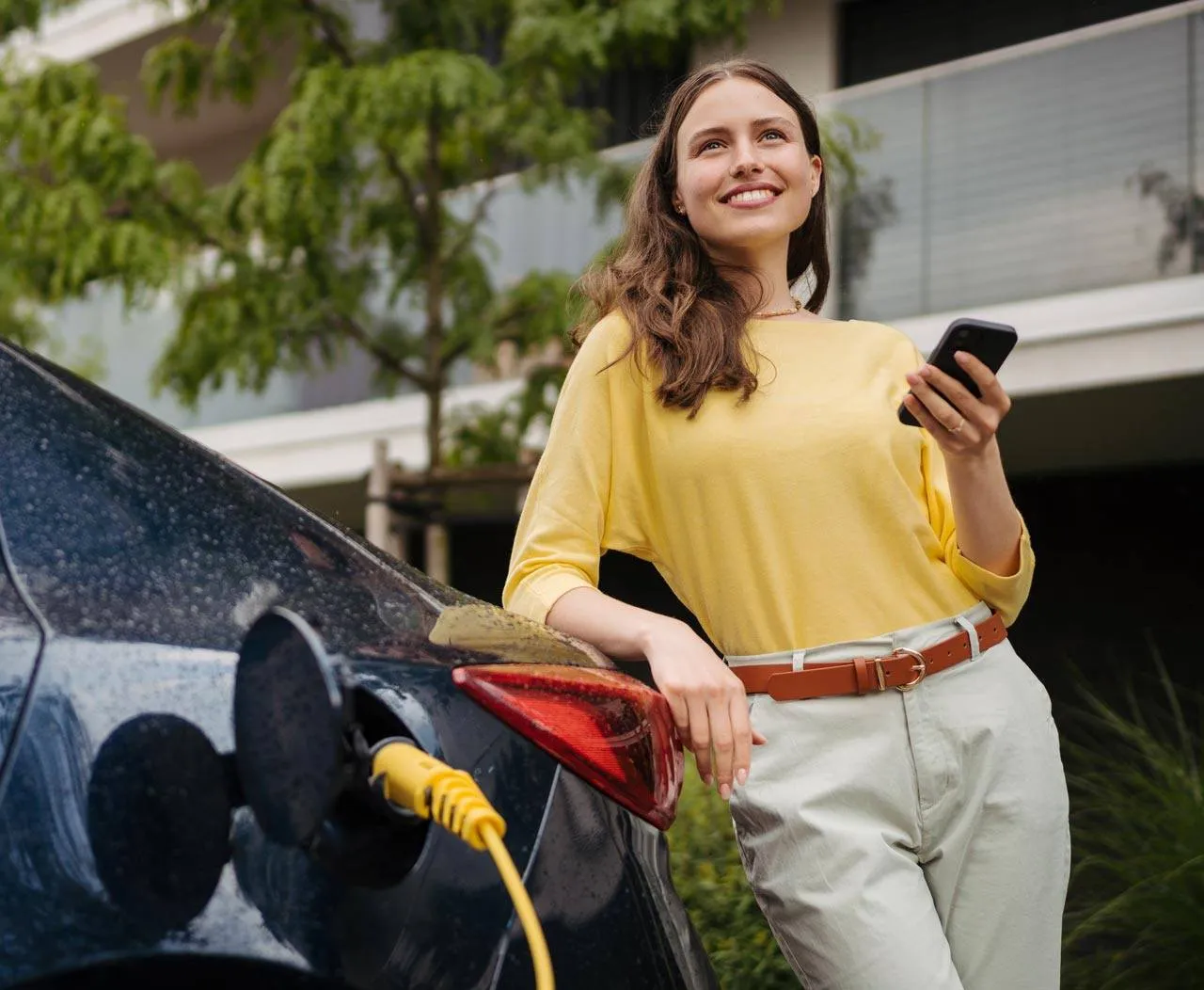 Woman leaning against an electric vehicle while it's charging with a phone in her hand.