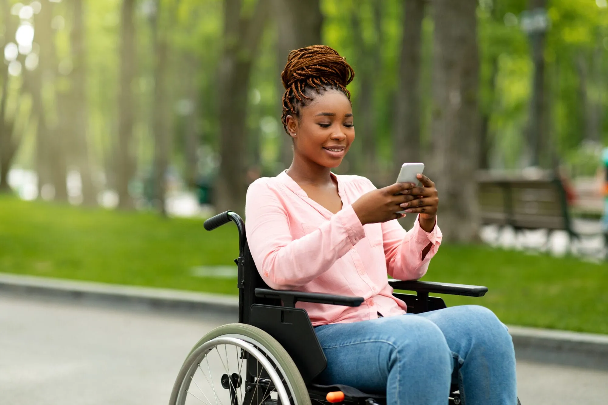 Happy handicapped black woman in wheelchair using smartphone, checking messages, browsing internet at city park