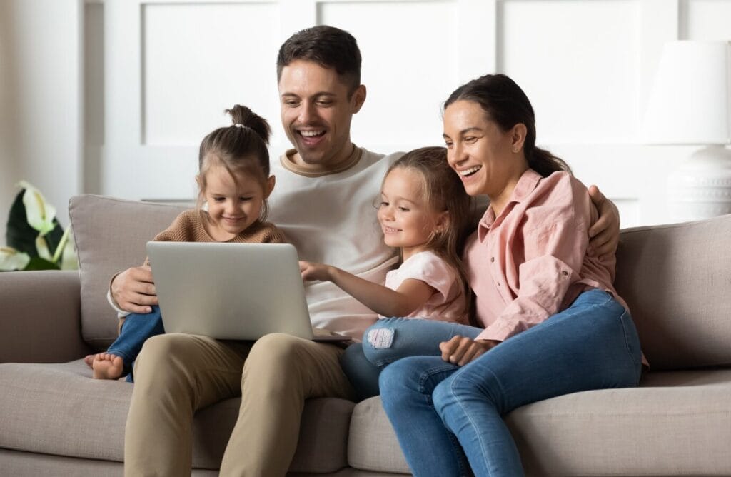 Family of father, mother, and two daughters sitting on couch looking at laptop